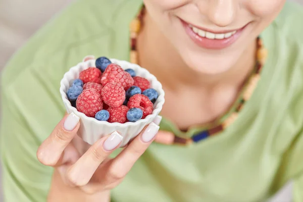 Vista recortada de mujer vegetariana sonriente sosteniendo tazón con frambuesas maduras frescas y arándanos - foto de stock