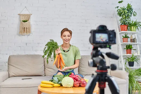 Vegetarian video blog, woman with fresh carrots near plant-based food in front of digital camera — Stock Photo