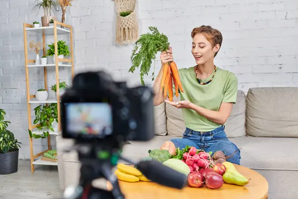 Joyful female video blogger with carrots near fresh plant-based food and blurred digital camera — Stock Photo