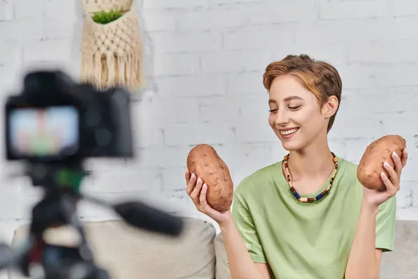 Alegre vegetariana mujer sosteniendo camote en frente de borrosa cámara digital durante video blog - foto de stock