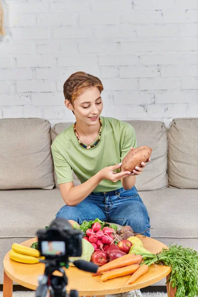 Smiley woman with sweet potato near fresh vegetables and fruits in front of blurred digital camera — Stock Photo