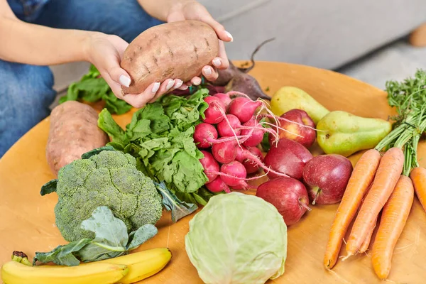 Vista cortada da mulher segurando batata-doce sobre pilha de vários legumes e frutas, vegetarianismo — Fotografia de Stock