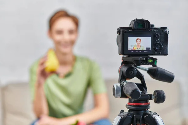 Selective focus on digital camera near blurred woman with ripe pier, video blog on plant-based diet — Stock Photo