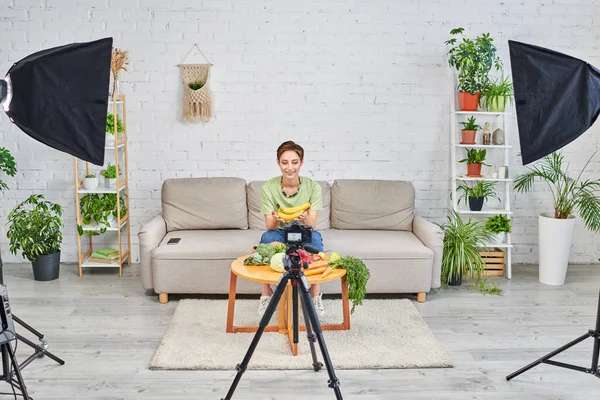 Женщина возле растительной пищи и цифровой камеры с софтбокс-ламами в гостиной, вегетарианский блог — стоковое фото