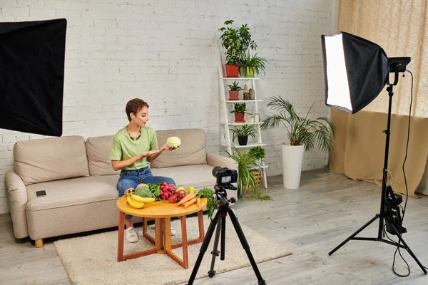 Вегетарианский блог, женщина с капустой возле стола с овощами и фруктами перед цифровой камерой — стоковое фото