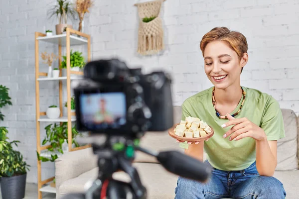 Happy woman with plate of tofu cheese near blurred digital camera, vlog on plant-based diets — Stock Photo