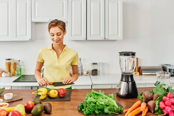Happy woman with knife near vegetables with fruits and electric blender in kitchen, plant-based diet — Stock Photo