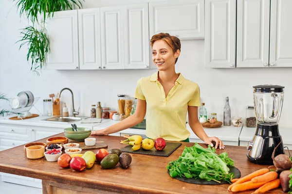 Радісна жінка біля столу з різними інгредієнтами рослинного походження та електричним блендером на сучасній кухні — стокове фото