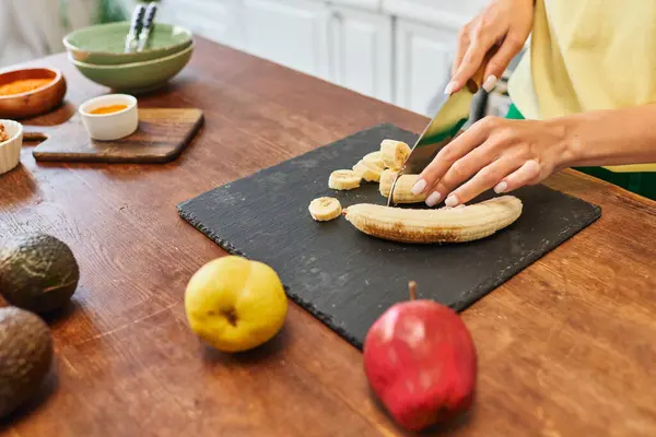Abgeschnittene Ansicht einer Frau, die reife Bananen in der Nähe von Äpfeln und Avocado schneidet, während sie eine vegetarische Mahlzeit zubereitet — Stockfoto