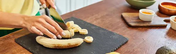 Vista cortada da mulher cortando banana fresca perto de ingredientes vegetarianos na mesa na cozinha, banner — Fotografia de Stock