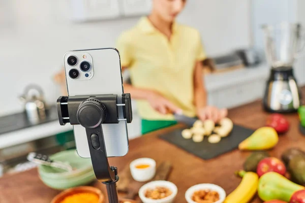Happy woman cutting banana during video blog near plant origin ingredients and smartphone in kitchen — Stock Photo