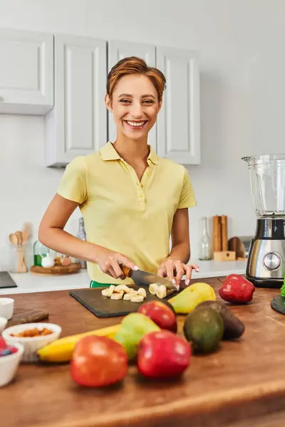 Mulher vegetariana feliz cortando banana madura perto de frutas frescas e liquidificador elétrico na cozinha moderna — Fotografia de Stock