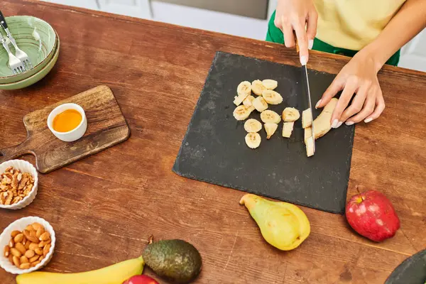 Vista recortada de la mujer cortando plátano cerca de frutas frescas y frutos secos en la cocina, cocina casera vegetariana - foto de stock