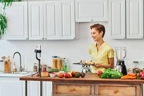 Happy woman with sliced banana on chopping board near smartphone and vegetarian ingredients, vlog — Stock Photo