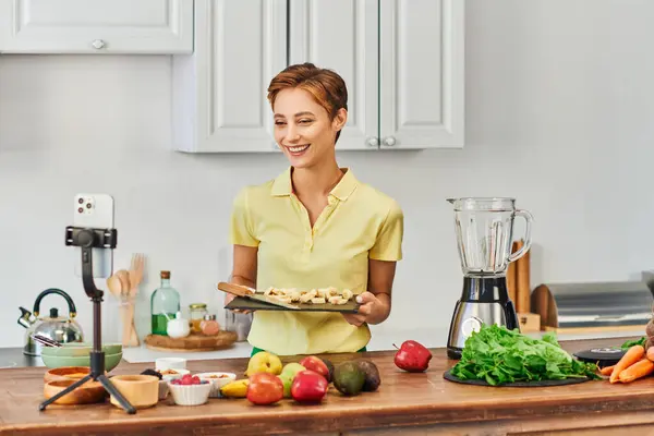 Woman with sliced banana on chopping board smiling during vegetarian culinary video blog in kitchen — Stock Photo
