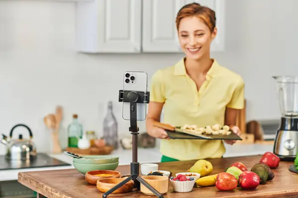 Smartphone em tripé perto de mulher com tábua de corte e frutas frescas com legumes na cozinha — Fotografia de Stock