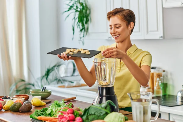 Mulher alegre com banana fatiada na tábua de corte perto do liquidificador e ingredientes vegetarianos na cozinha — Fotografia de Stock