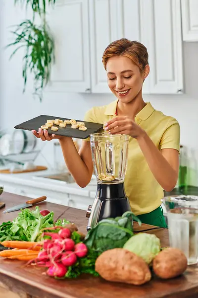 Heureux végétarien femme mettre des tranches de banane dans mélangeur électrique près de légumes frais dans la cuisine — Photo de stock