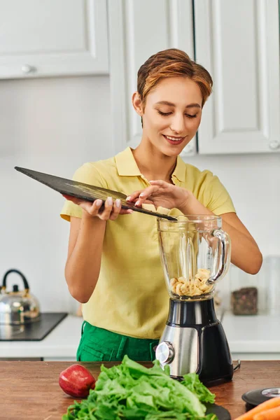 Smiling woman putting sliced banana into electric blender near fresh lettuce, plant-focused diet — Stock Photo