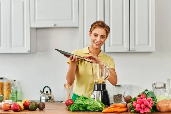 Smiley woman with chopping board near electric blender and fresh fruits with vegetables in kitchen — Stock Photo