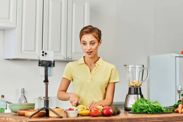 Женщина режет фрукты рядом со смартфоном на треноге и электрический блендер дома, растительный диетический блог — стоковое фото
