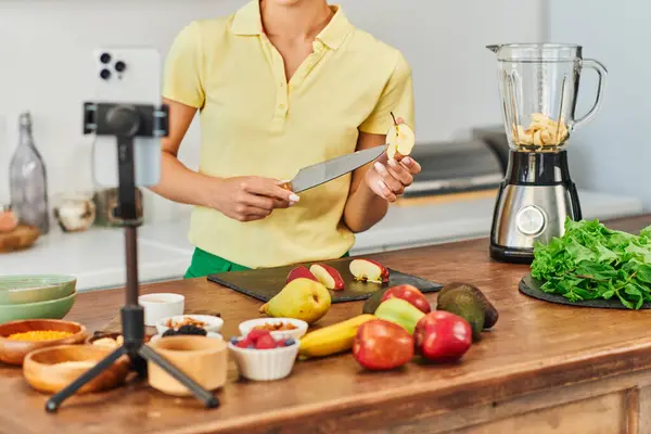 Обрізаний вид на вегетаріанку з ножем і розрізаним яблуком біля смартфона і фруктів на кухні — стокове фото