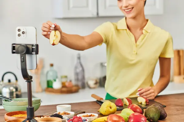 Обрезанная улыбающаяся женщина держит вырезанное яблоко возле смартфона на штативе и свежие фрукты на кухне — стоковое фото