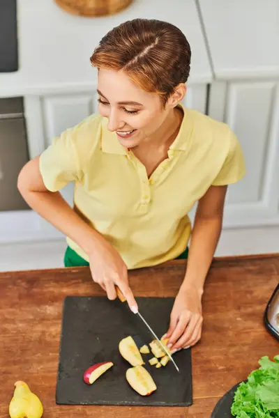 Mulher vegetariana alegre cortando maçã madura e preparando refeição saudável na cozinha, vista de alto ângulo — Fotografia de Stock