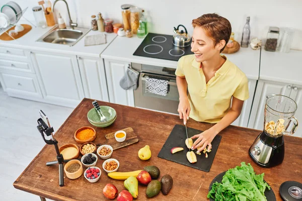 Весела жінка, що ріже фрукти біля електричного блендера та смартфона на тринозі, вегетаріанський відео блог — стокове фото