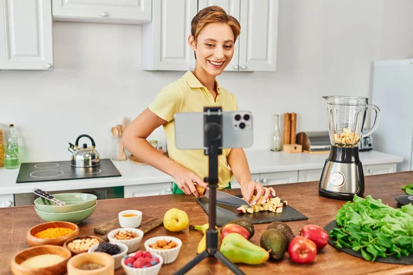Happy vegetarian woman cutting fruits near blender and smartphone on tripod, plant-based diets vlog — Stock Photo