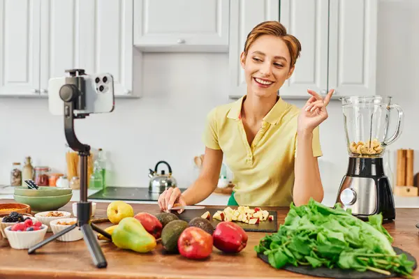 Веселая вегетарианка смотрит на смартфон на штативе рядом фрукты и электрический блендер, блог — стоковое фото