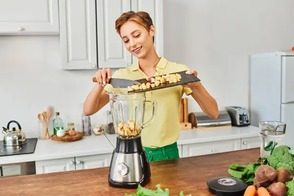 Sonriente mujer añadiendo manzana picada en licuadora eléctrica cerca de ingrediente de origen vegetal, dieta vegetariana - foto de stock