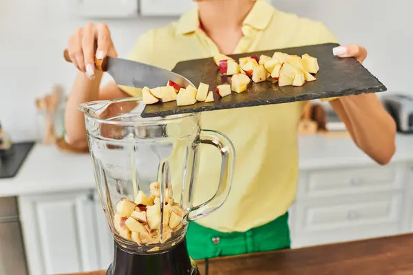 Femme végétarienne avec planche à découper et couteau ajoutant pomme hachée dans mélangeur électrique, vue recadrée — Photo de stock