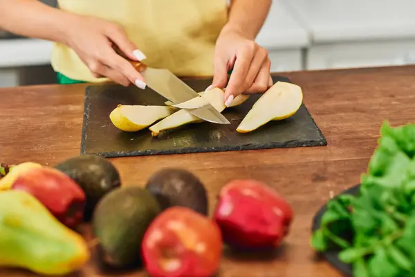 Vista cultivada de mulher vegetariana cortando pêra madura perto de frutas frescas na cozinha, dieta focada em plantas — Fotografia de Stock