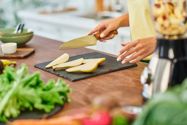 Vista ritagliata della donna vegetariana con coltello vicino alla pera affettata e lattuga sfocata in cucina — Foto stock