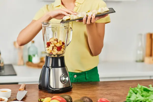 Vista cortada da mulher adicionando frutas picadas no liquidificador elétrico, cozinha caseira vegetariana saudável — Fotografia de Stock