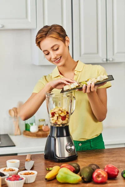 Mulher alegre adicionando frutas picadas no liquidificador elétrico enquanto cozinha, receita vegetariana — Fotografia de Stock