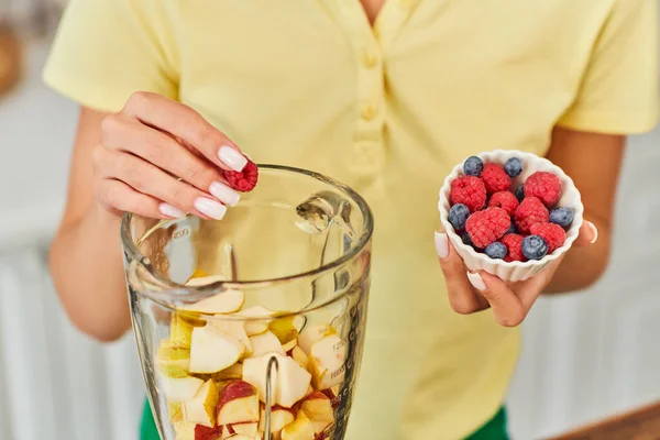 Cropped view of vegetarian woman adding raspberries and blueberries into blender with chopped fruits — Stock Photo