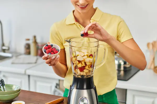 Обрезанный вид улыбающейся женщины, добавляющей ягоды в электрический блендер с нарезанными фруктами, вегетарианская диета — стоковое фото