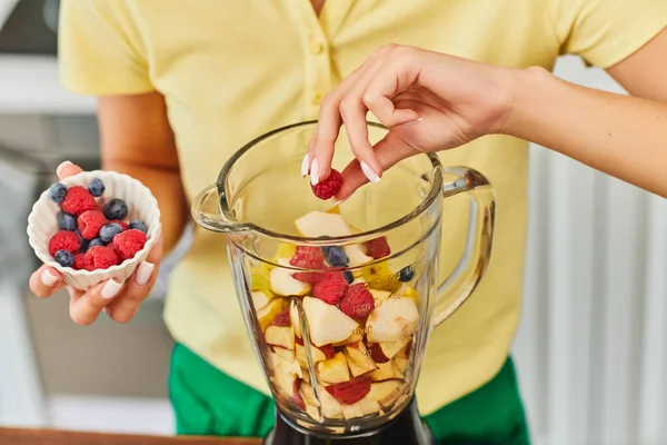 Cropped woman adding raspberries and blueberries into blender with chopped fruits, vegetarian diet — Stock Photo