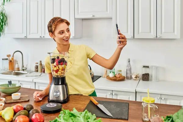 Joyful vegetarian woman with smartphone taking selfie near blender with chopped fruits in kitchen — Stock Photo