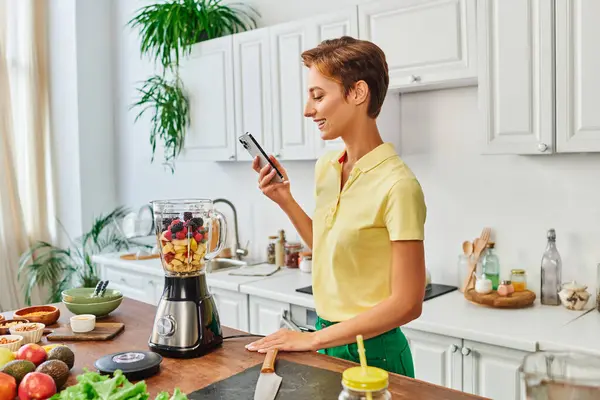Mulher feliz tirar foto de liquidificador elétrico com frutas picadas na cozinha moderna, vegetarianismo — Fotografia de Stock