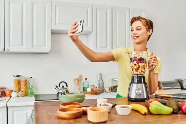 Mulher vegetariana satisfeito tomar selfie no smartphone perto do liquidificador elétrico com frutas frescas picadas — Fotografia de Stock