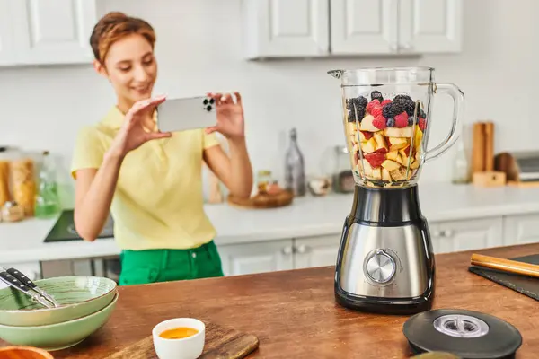 Happy woman with smartphone taking photo of electric blender with chopped fruits, vegetarian recipe — Stock Photo