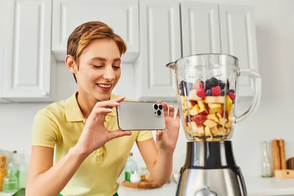 Mujer positiva tomando fotos de licuadora eléctrica con frutas picadas, deliciosa dieta a base de plantas - foto de stock