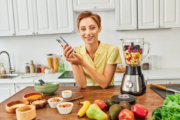 Mujer vegetariana con teléfono inteligente sonriendo a la cámara cerca de origen vegetal alimentos y licuadora con frutas - foto de stock