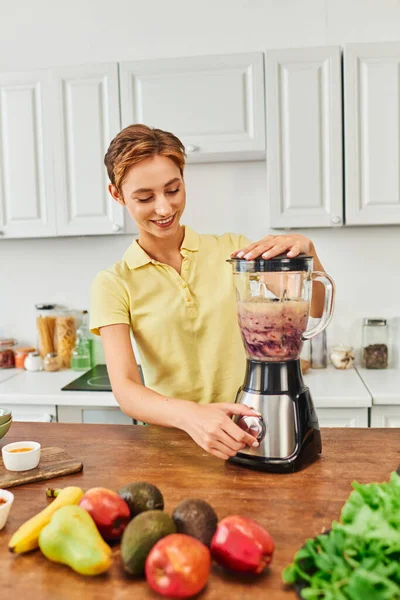 Happy woman preparing delicious smoothie while grinding fresh fruits in kitchen, vegetarian recipe — Stock Photo