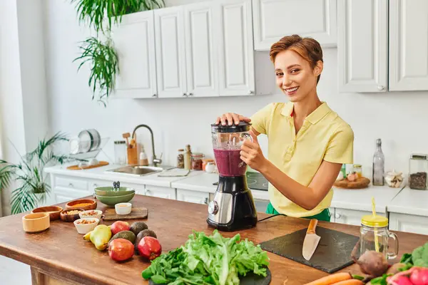 Vegetarian woman preparing smoothie in electric blender near fresh fruits and vegetables in kitchen — Stock Photo