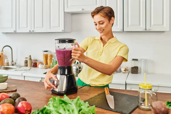 Smiley woman preparing vegetarian smoothie in electric blender near fresh fruits and vegetables — Stock Photo