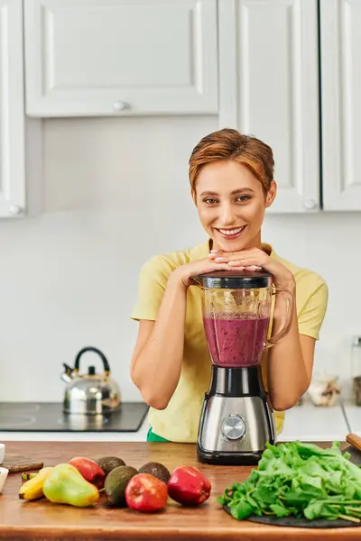 Mulher vegetariana sorrindo para a câmera perto do liquidificador com smoothie saudável na cozinha, dieta à base de plantas — Fotografia de Stock
